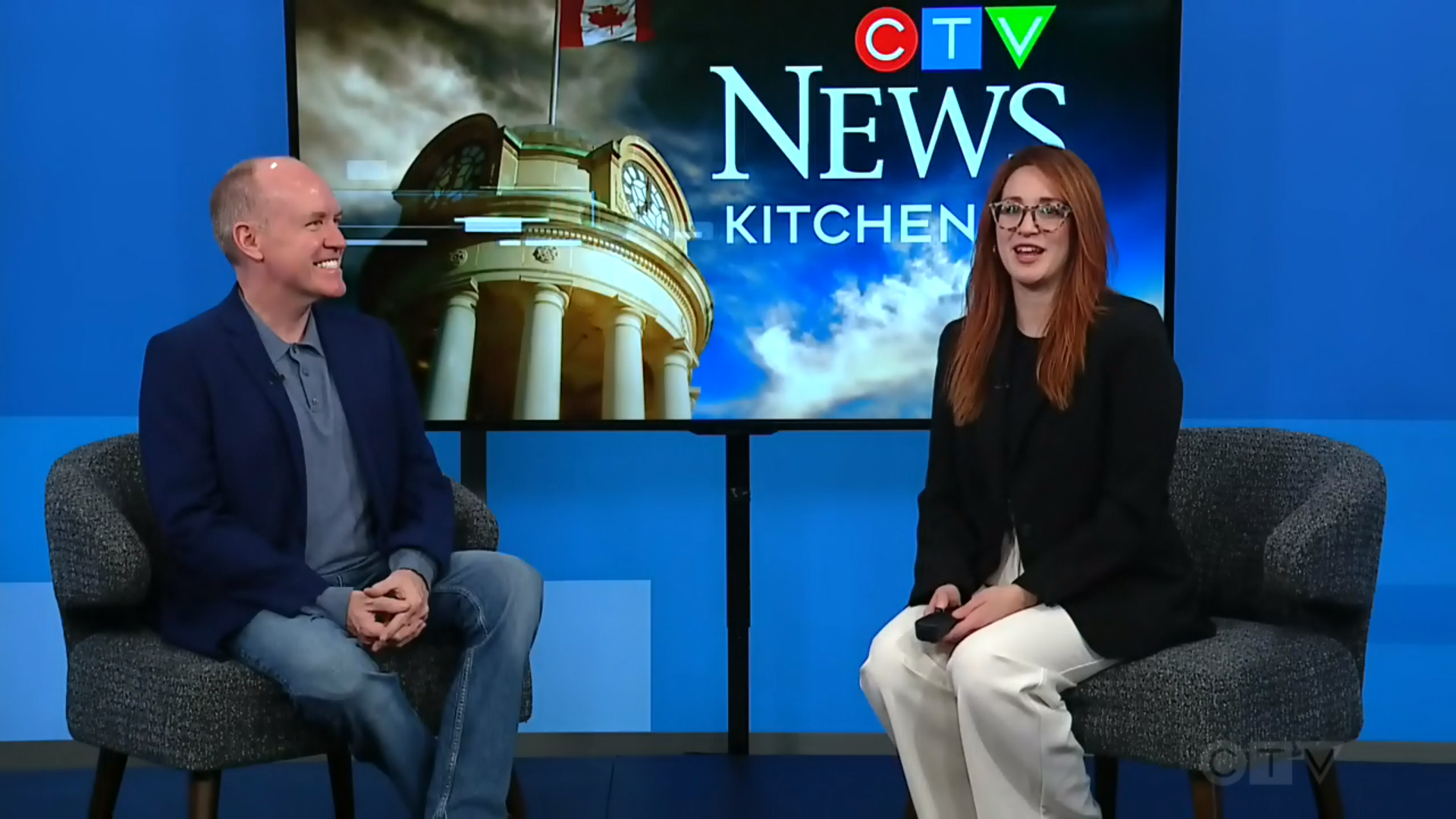 David Connolly and Daryl Morris sitting in front of a CTV News Kitchener backdrop. David wears a dark blue shirt and jeans, while Daryl on the right is dressed in a black blazer and white pants. Both are seated on grey upholstered chairs. The backdrop features the CTV News Kitchener logo, a clock tower, and the Canadian flag.